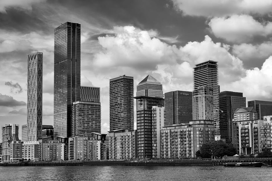 A tall modern office building with a reflective glass facade stands prominently against a partly cloudy sky, surrounded by other high-rise structures with various architectural styles, including one w