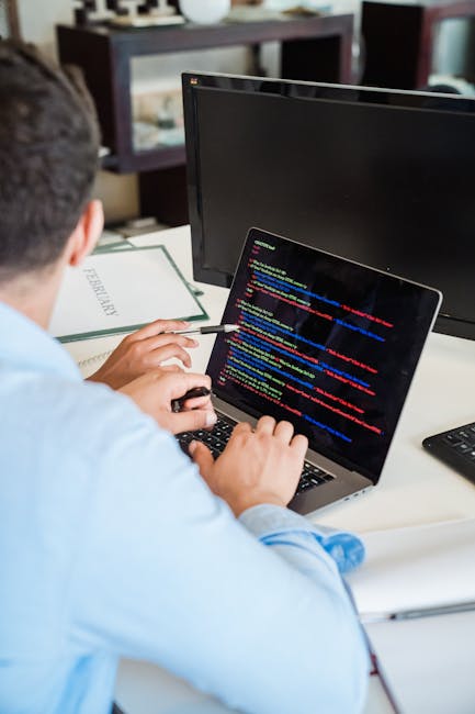 A person seated at a desk working on a laptop displaying lines of colorful computer code, with an external monitor positioned in front of them. The individual, wearing a light blue shirt, is using a m