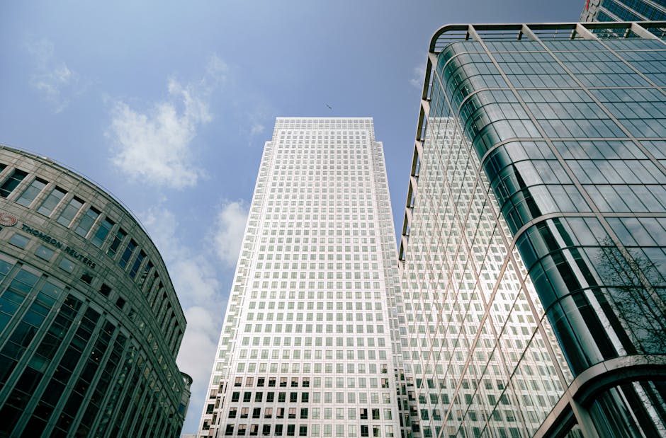 A tall, modern commercial office building with a curved corner design, constructed with a combination of glass and steel panels. The glass façade reflects the clear blue sky, and the windows are fram